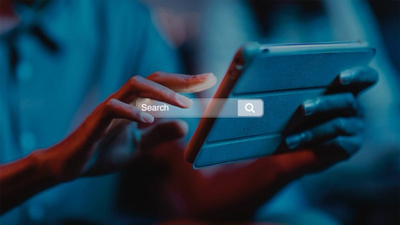 Una mano sfiora una barra di ricerca luminosa davanti a un tablet, in luce blu e rossa
