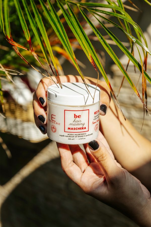 Una mano tiene un vasetto di crema Essere Bio fra le foglie di palma al sole