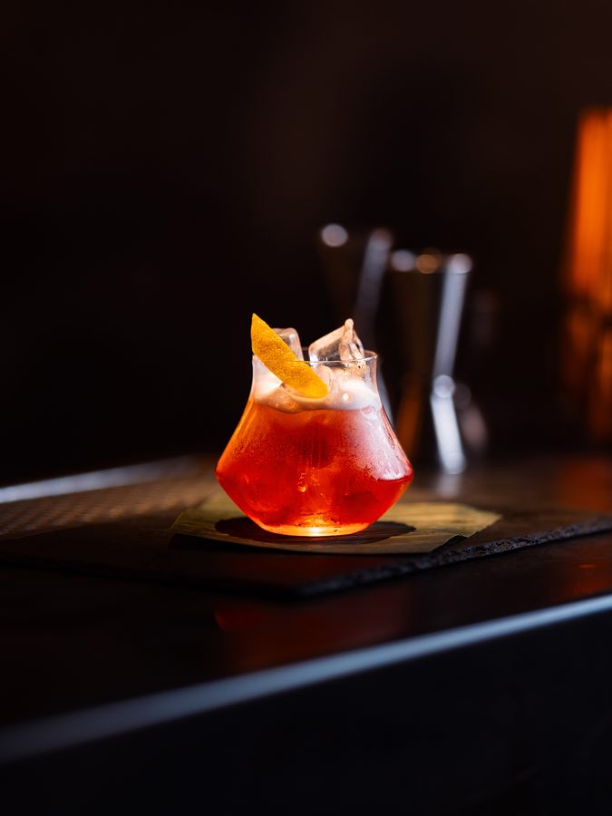 Un cocktail arancio con la buccia, sul bancone scuro di MAG 56