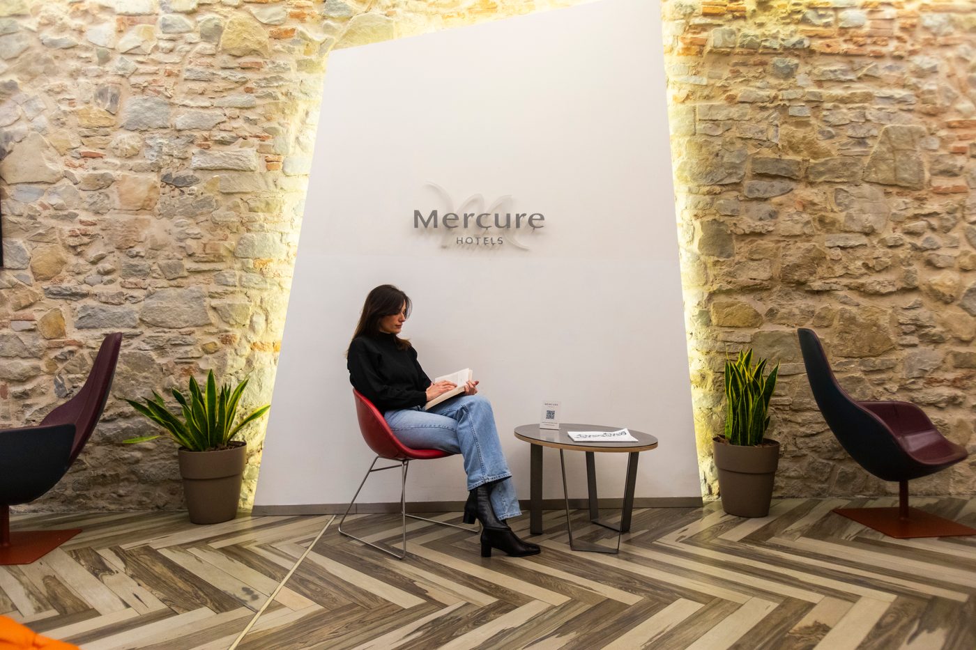 Una ragazza legge in poltrona accanto al muro di pietra col logo Mercure